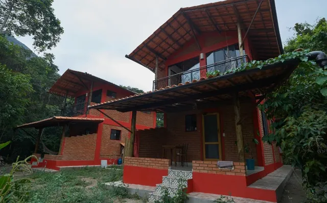 Casa de tijolos com telhado de telha e varanda, cercada de mata